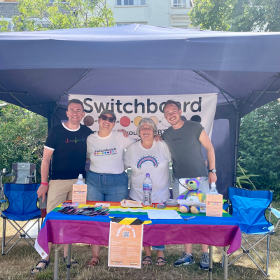 Image of four white people underneath a gazebo smiling. Behind them is the Switchboard banner and infront is a table with a rainbow flag