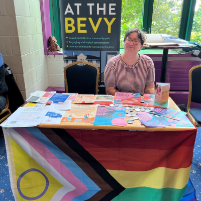 Photo of a white non-binary person sitting at a table with a progress flag hanging below and flyers and other printed materials on the table