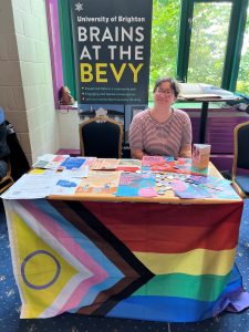 Photo of a white non-binary person sitting at a table with a progress flag hanging below and flyers and other printed materials on the table