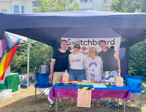 Image of four white people underneath a gazebo smiling. Behind them is the Switchboard banner and infront is a table with a rainbow flag