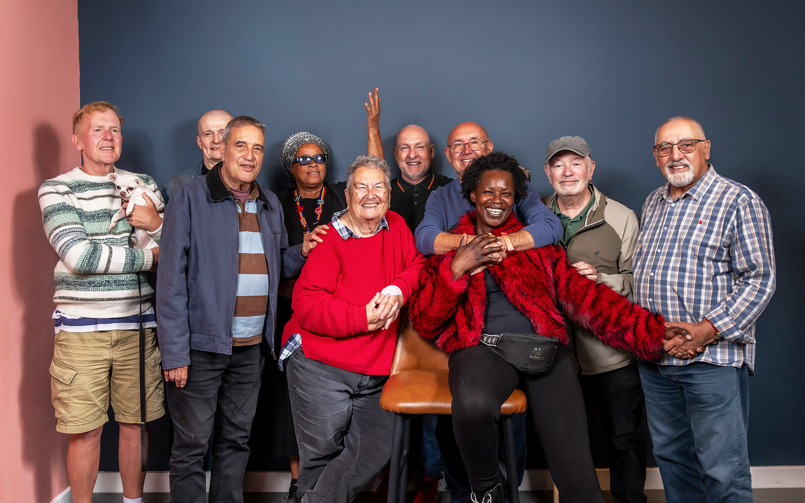 Group portrait of older LGBTQ+ community members smiling together against a dark blue studio backdrop, with one person seated at the front and others standing around them.