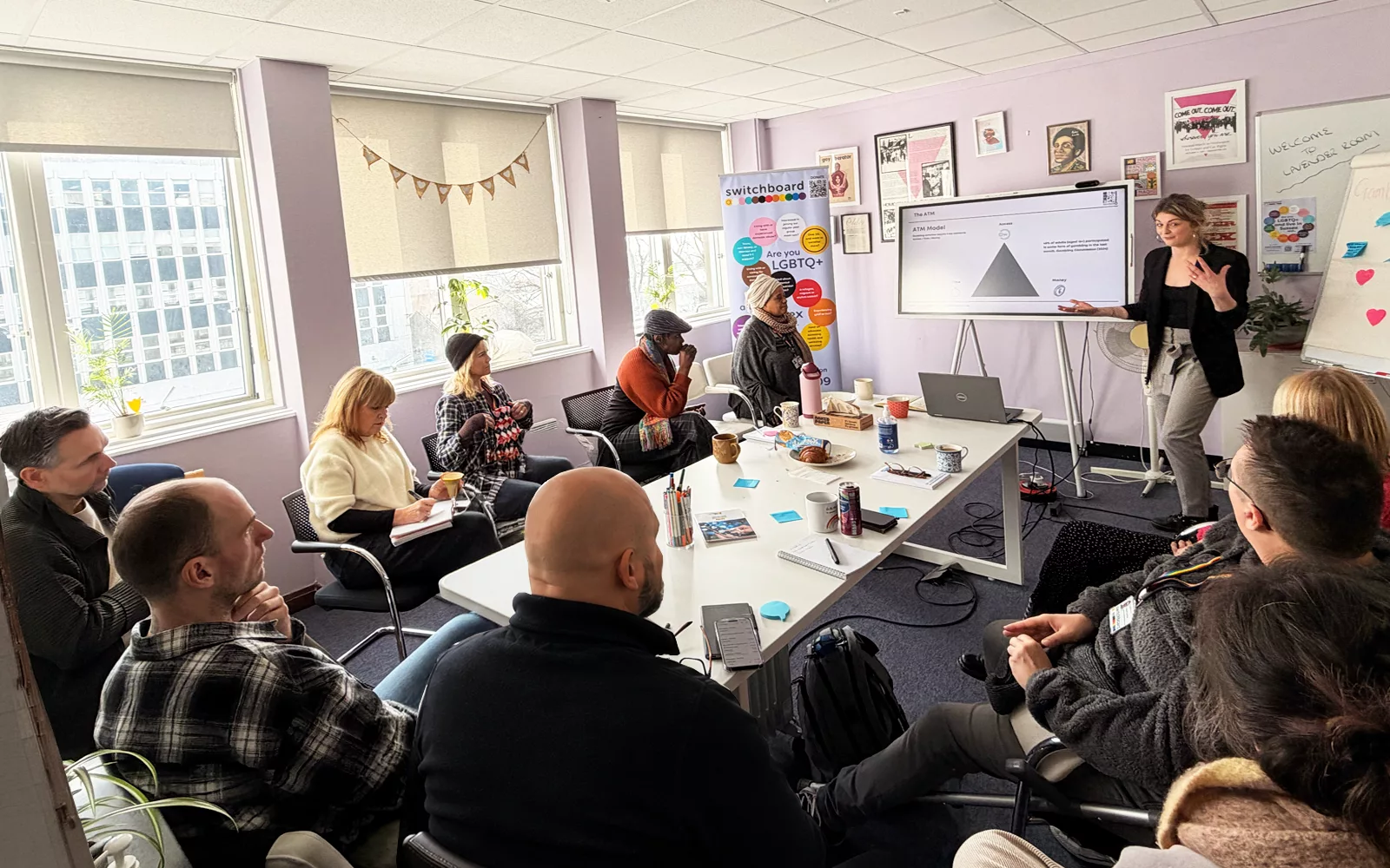 People seated around a meeting room table during an LGBTQ+ inclusion workshop, with a presenter standing beside a screen and Switchboard training materials visible in the room.